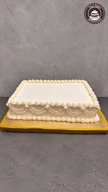 Elegant white rectangular celebration cake featuring intricate vintage piping, scalloped borders, pearl details, and a gold cake board
