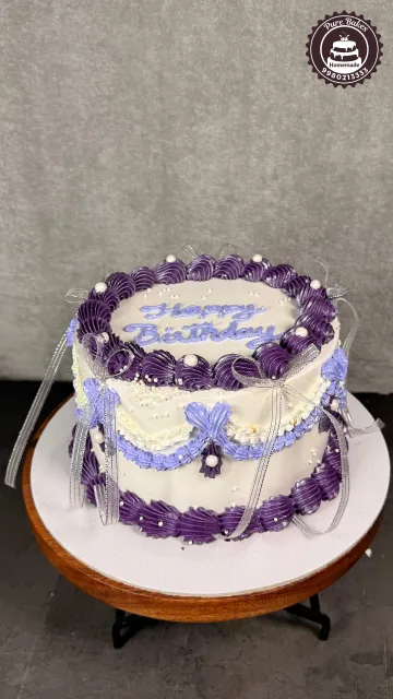 Purple and white vintage birthday cake decorated with piped buttercream borders, ribbons, pearls, and Happy Birthday lettering